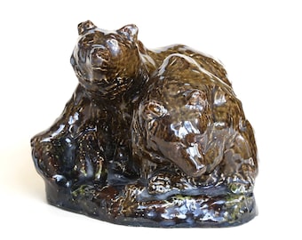 May include: A ceramic sculpture of two brown bears, side-by-side, resting on a textured base. The bears have a glossy finish, with visible details in their fur and facial features. The sculpture is a decorative item.