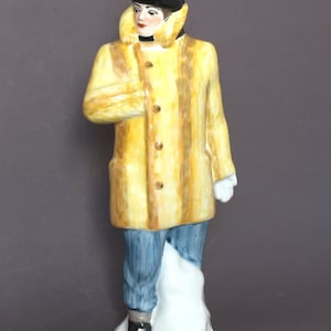 Puede incluir: Una figurita de cerámica de una persona que lleva un abrigo amarillo con cuello de piel, un sombrero negro y pantalones azules. La figurita está de pie sobre una base blanca.
