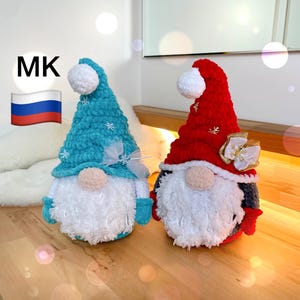 May include: Two plush gnome figures. One is teal with white accents, the other is red with gold accents. Both have white beards, tan noses, and white pom-poms on their hats. The gnomes are decorated with snowflakes and bows.