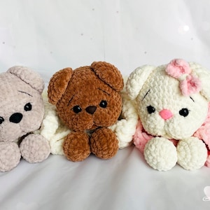 May include: Three handmade crochet stuffed animals in a row. The first is a light beige bear, the second is a brown bear, and the third is a white bunny with a pink bow. All are lying down on a white surface.