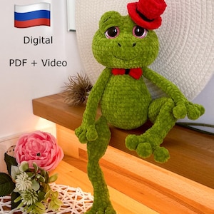 May include: Green crocheted frog wearing a red hat and a red bow tie. The frog is sitting on a wooden surface. Digital PDF + Video.