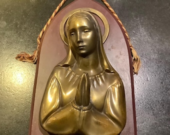 May include: A gold-toned metal sculpture of a woman with her hands clasped in prayer. The sculpture is mounted on a dark wood plaque with a rope hanger.