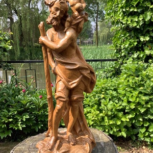 Könnte beinhalten: Eine Holzskulptur des Heiligen Christophorus, der ein Kind auf seiner Schulter trägt. Die Skulptur ist realistisch gearbeitet und zeigt aufwendige Details. Der Heilige hält einen Stab in seiner linken Hand und das Kind hält einen kleinen Gegenstand in seiner rechten Hand.