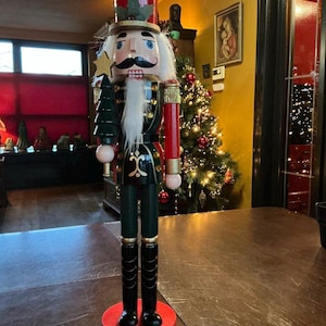 Può includere: Una statuina schiaccianoci in legno con un design tradizionale da soldato. La statuina ha un cappello rosso, una barba bianca e dei baffi neri. Tiene in mano un albero verde e una stella d'oro. Lo schiaccianoci è su una base rossa.