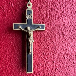 Könnte beinhalten: Ein Kruzifix mit einer goldfarbenen Jesusfigur und einem schwarzen Kreuz mit goldenen Verzierungen. Das Kreuz hängt vor einer strukturierten roten Wand. Auf dem oberen Teil des Kreuzes stehen die Buchstaben INRI.
