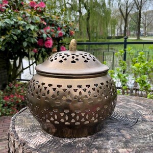 Op de afbeelding: Een bronzen wierookbrander met een decoratief, ingewikkeld ontwerp. De brander heeft een deksel met een kleine knop bovenop en staat op een boomstronk.