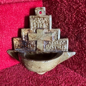 Puede incluir: Una pila de agua bendita de cerámica beige con las palabras "SAINT" y "PARIS" inscritas. La pila tiene un diseño escalonado con una pequeña cuenca en la parte inferior y un agujero en la parte superior para colgar. El fondo es una tela roja texturizada.