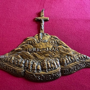 Puede incluir: Placa religiosa de color bronce con una cruz en la parte superior y texto. La placa presenta las palabras "1933 APRIL 1934 HEILIG JAAR" y "JEZUSWEESKONING OVER ONS OVERONSVOLK" en una fuente decorativa. La placa está sobre un fondo magenta.