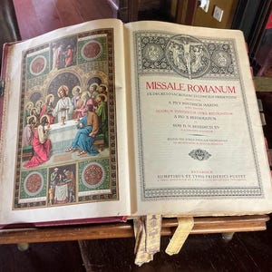 May include: An open antique book, titled "MISSALE ROMANUM," with ornate illustrations and text. The pages display religious artwork depicting figures at a table. The book is propped open on a wooden stand with metal clasps.