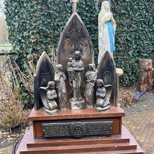 Op de afbeelding: Een religieus beeld met een centrale figuur die een kind vasthoudt, geflankeerd door andere figuren en engelen. Het beeld staat op een houten voet met een plaquette. Een kruis bekroont de gewelfde structuur. Het kleurenpalet is voornamelijk brons en bruin.