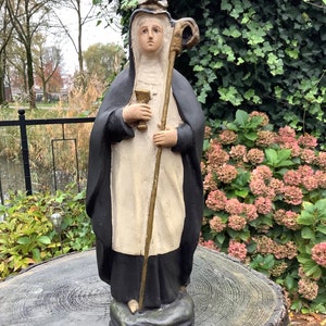 Puede incluir: Una estatua en blanco y negro de una santa con una corona dorada y un bastón. La estatua está de pie sobre una base de madera con la inscripción "S'ADELE".