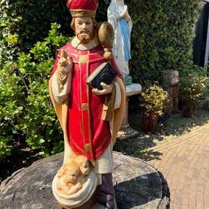 Könnte beinhalten: Detaillierte Statue einer religiösen Figur, die ein rotes und goldenes Gewand und eine hohe rote Mütze trägt. Die Figur hält ein Buch und einen Stab, mit zwei Engeln an der Basis. Im Hintergrund eine Statue der Jungfrau Maria.
