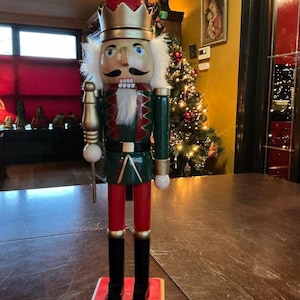 Puede incluir: Una figura de cascanueces de madera, de pie sobre una base roja. La figura presenta un sombrero rojo, una corona dorada y un uniforme verde y rojo. Tiene barba y bigote blancos y sostiene un bastón dorado. Un árbol de Navidad está al fondo.