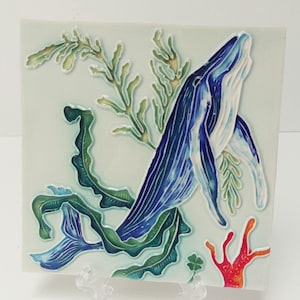 Puede incluir: Azulejo de cerámica cuadrado con una ballena azul rompiendo la superficie, rodeada de algas verdes y amarillas y coral rojo. La ballena tiene marcas azules y blancas detalladas. El fondo es azul claro.