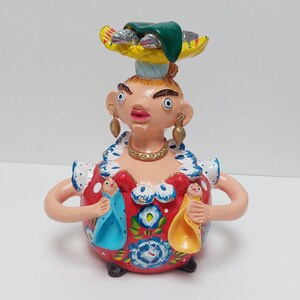 May include: A colorful ceramic figurine of a woman wearing a red dress with floral designs and carrying two smaller figures on her arms. She is wearing a green hat with fish on top and gold earrings.