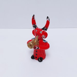 May include: A red clay figurine of a devil playing a gold trumpet. The devil has black horns and a black and white striped tail. The figurine is about 5 inches tall.