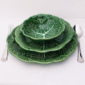 May include: A set of three green ceramic plates and a bowl, all shaped like cabbage leaves. The plates are stacked on top of each other, with the bowl on top of the plates.