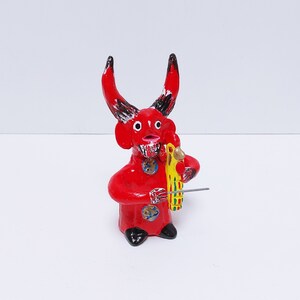 May include: Red clay figurine of a devil playing a musical instrument. The devil has black horns and a red and black patterned body. The instrument is yellow and has a red and green design.