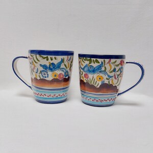 May include: Two ceramic mugs with blue and white floral designs and blue birds. The mugs have brown, orange, and yellow accents.