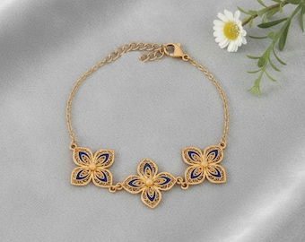 Op de afbeelding: Goudkleurige armband met drie bloemvormige filigraan bedels met blauwe emaille details. De armband heeft een fijne ketting en een karabijnsluiting. Een madeliefje en groen zijn op de achtergrond.