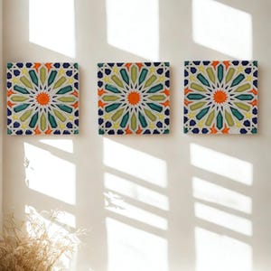 May include: Three square decorative tiles with a geometric starburst design. The tiles feature a central orange circle surrounded by teal, yellow, and green shapes, with a dark blue border. The tiles are mounted on a white wall.