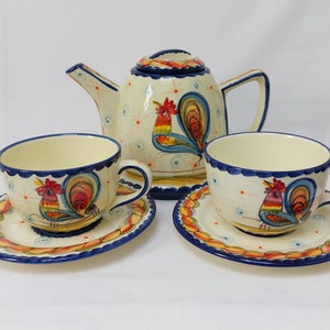 Può includere: Un set da tè in ceramica con una teiera e due tazze e piattini. Il set presenta un design a gallo con accenti blu, gialli, rossi e verdi su uno sfondo bianco.