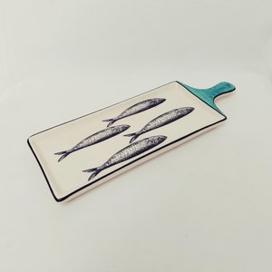 May include: A white rectangular serving dish with a teal handle. The dish is decorated with a black and white illustration of five fish.