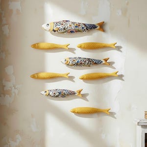 Könnte beinhalten: Wanddekoration mit Keramikfischen. Einige Fische sind gelb, andere haben aufwendige Blumenmuster in Blau, Weiß und Rot. Die Fische sind an einer strukturierten, cremefarbenen Wand befestigt.