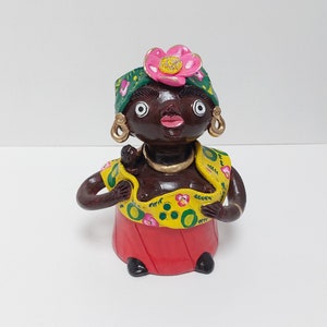 May include: A ceramic figurine of a woman with dark brown skin, wearing a green and yellow patterned dress with a pink flower on her head. She is holding a small child in her arms.