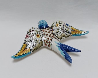 May include: A ceramic swallow figurine with blue, yellow and green floral patterns. The swallow is in flight with its wings spread.
