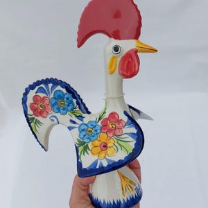 Puede incluir: Una figura de gallo de cerámica blanca con cresta y barbilla rojas. El gallo está decorado con motivos florales azules y blancos y tiene un pico amarillo. La figura está de pie sobre una base blanca con detalles azules y blancos.