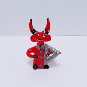 May include: A red figurine of a devil-like creature with black-tipped horns, a blue bow tie, and a silver triangle-shaped instrument. The figure has a smiling expression and is wearing a red coat with blue buttons. The figurine is standing on a white surface.