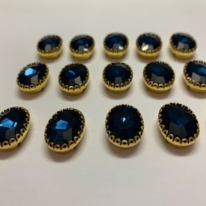 Könnte beinhalten: Eine Ansammlung ovaler, dunkelblauer Edelstein-Knöpfe mit goldfarbenen Metallsockeln und dekorativen Rändern. Die Knöpfe sind in Reihen angeordnet und zeigen ihre facettierten Oberflächen und reflektierenden Eigenschaften.