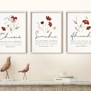 Puede incluir: Tres impresiones artísticas enmarcadas con diseños florales en acuarela y caligrafía. Cada impresión presenta una palabra diferente: "Chesed", "Simcha" y "Ahavah". Las impresiones se exhiben sobre un mueble blanco con esculturas decorativas de pájaros.