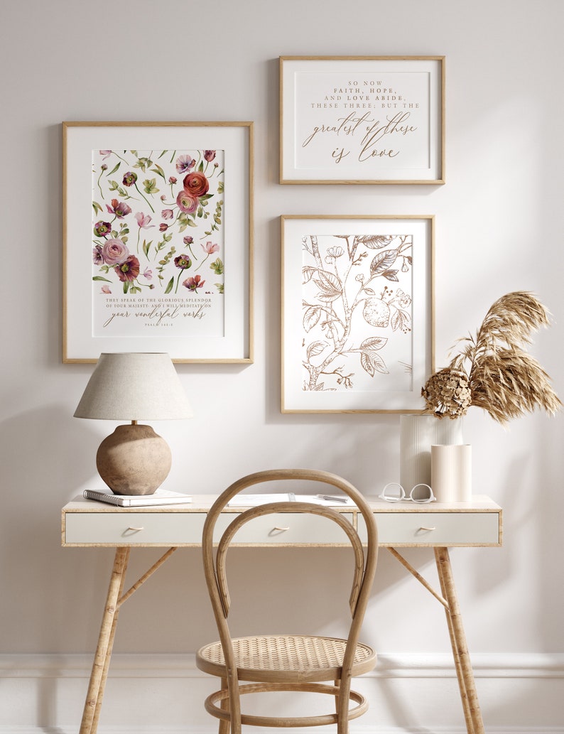 May include: Three framed prints with floral designs and inspirational quotes. The prints are hung on a white wall above a white desk with a wicker chair and a lamp. The quote on the top print reads "So now faith, hope, and love abide these three: but the greatest of these is love."
