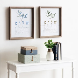 May include: Two framed prints featuring a watercolour illustration of olive branches and the Hebrew word "Shalom" in blue and gold lettering. The print on the left reads "Shalom is the name of the Holy One, the name of Israel, the name of the Messiah, a manifestation of Divine Grace." The print on the right reads "Shalom is a blessing, a manifestation of Divine Grace, Peace, Prosperity, Completeness, Perfection, Tranquility, Health, Prosperity, Salvation, Harmony, Security, Friendship, Fulfillment."