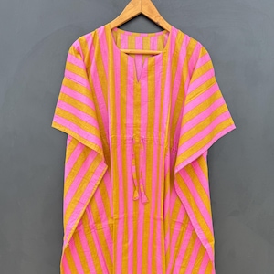 May include: A pink and gold striped caftan dress hanging on a wooden hanger. The dress has a V-neckline and a drawstring waist. The fabric is lightweight and appears to be cotton or a similar material.