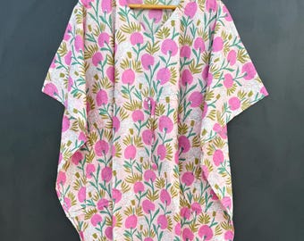 May include: A pink, white, and green floral print kaftan dress. The dress features a V-neck and short sleeves. The floral pattern includes large pink flower shapes with green stems and leaves. The dress is hanging on a wooden hanger.