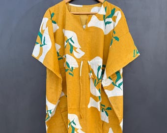 May include: Mustard yellow caftan dress with a white floral pattern and green accents. The dress has a V-neck, short sleeves, and a drawstring waist. It is hanging on a wooden hanger against a dark gray background.