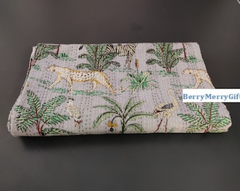 May include: A gray kantha quilt with a jungle animal print. The quilt features a leopard, zebra, flamingo, and palm trees. The quilt is folded and lying on a dark gray surface.