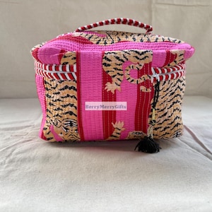 Puede incluir: Un neceser acolchado con un estampado de rayas de tigre en tonos rosa y beige. El bolso tiene un asa y una borla negra. El bolso es rectangular con cierre de cremallera.