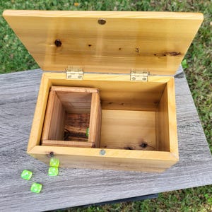 May include: Open wooden box with a hinged lid, revealing a smaller wooden box inside. The box is made of light-colored wood with visible grain. The lid is slightly ajar, and the box sits on a gray surface. Several small, translucent green cubes are scattered nearby.