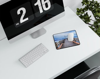 May include: A white computer keyboard and a white computer mouse are on a white desk with a black computer monitor and a mouse pad with a photo of a wooden pier extending into a body of water.