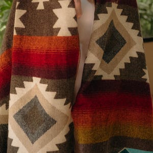May include: A woven blanket featuring a geometric pattern in shades of brown, cream, red, and teal. The textile displays repeating diamond and star motifs across its surface, with horizontal bands of color integrated into the overall design.
