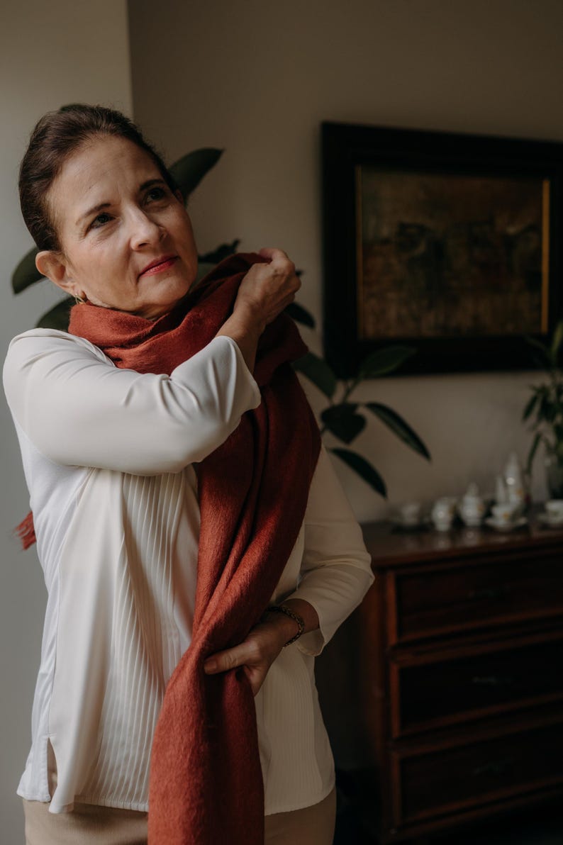 K&ouml;nnte beinhalten: Eine Frau tr&auml;gt eine wei&szlig;e Bluse und einen rostfarbenen Schal. Sie richtet den Schal um ihren Hals.