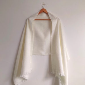 Puede incluir: Un chal rectangular de color blanco roto liso, hecho de una tela ligera y texturizada. La prenda presenta bordes rectos y detalles de flecos cortos y delicados a lo largo del dobladillo inferior, colgado en una percha de madera.