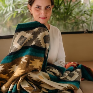 Może przedstawiać: Kobieta siedzi na kanapie i trzyma kolorowy koc z geometrycznym wzorem. Koc jest wykonany z wełny i ma miękką fakturę. Koc jest złożony na pół, a kobieta trzyma go na kolanach.