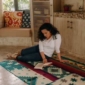 K&ouml;nnte beinhalten: Eine rechteckige Webdecke mit einem geometrischen Muster in Petrol, Creme, Dunkelrot und Grau. Das Textil zeigt wiederkehrende Rauten- und sternartige Formen auf der gesamten Oberfl&auml;che, mit einem durchgehenden dunkelroten Band in der Mitte.