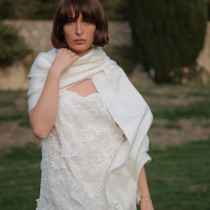 Puede incluir: Un vestido blanco sin mangas con un intrincado encaje floral superpuesto y un dobladillo festoneado, combinado con un chal rectangular blanco liso y suave drapeado sobre los hombros. El vestido presenta un escote recto y silueta ajustada.