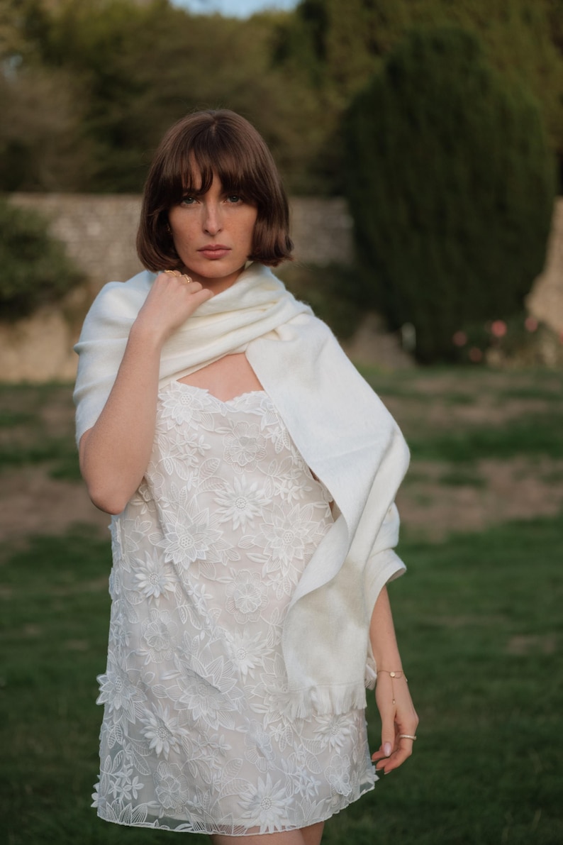 Puede incluir: Un vestido blanco sin mangas con un intrincado encaje floral superpuesto y un dobladillo festoneado, combinado con un chal rectangular blanco liso y suave drapeado sobre los hombros. El vestido presenta un escote recto y silueta ajustada.